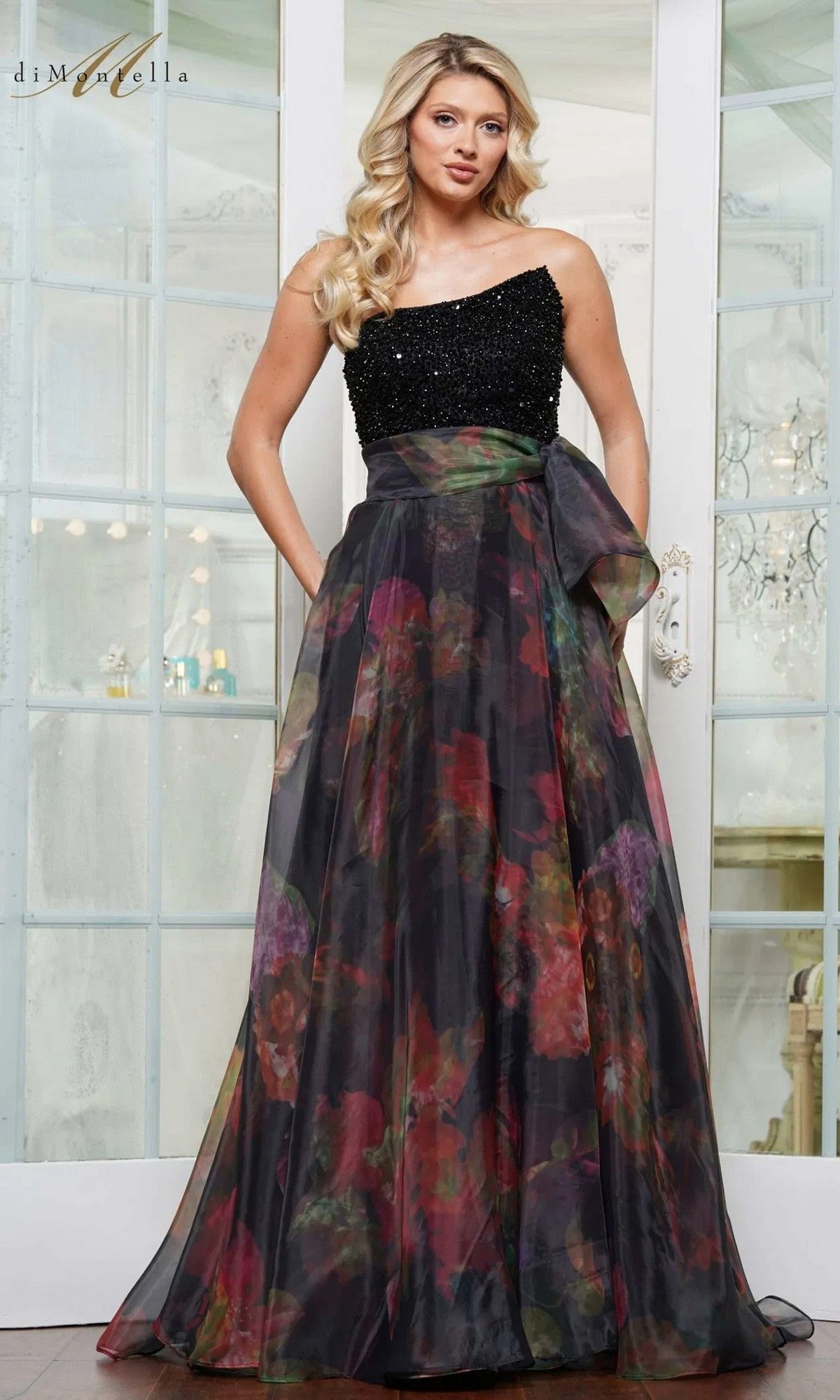 A woman poses gracefully indoors in front of glass-paneled doors, wearing the Long Formal Dress: Rina Di Montella RD3213 in black, featuring a beaded bodice top, floral-printed organza skirt, and a large side bow.